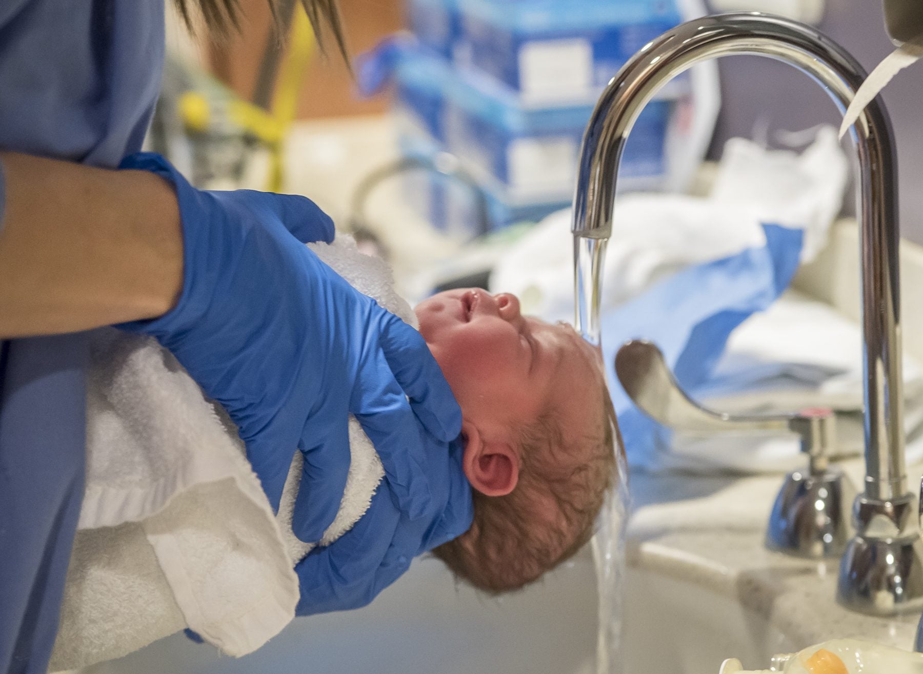 A nurse in blue gloves holds a newborn under a running tap for the first bath.