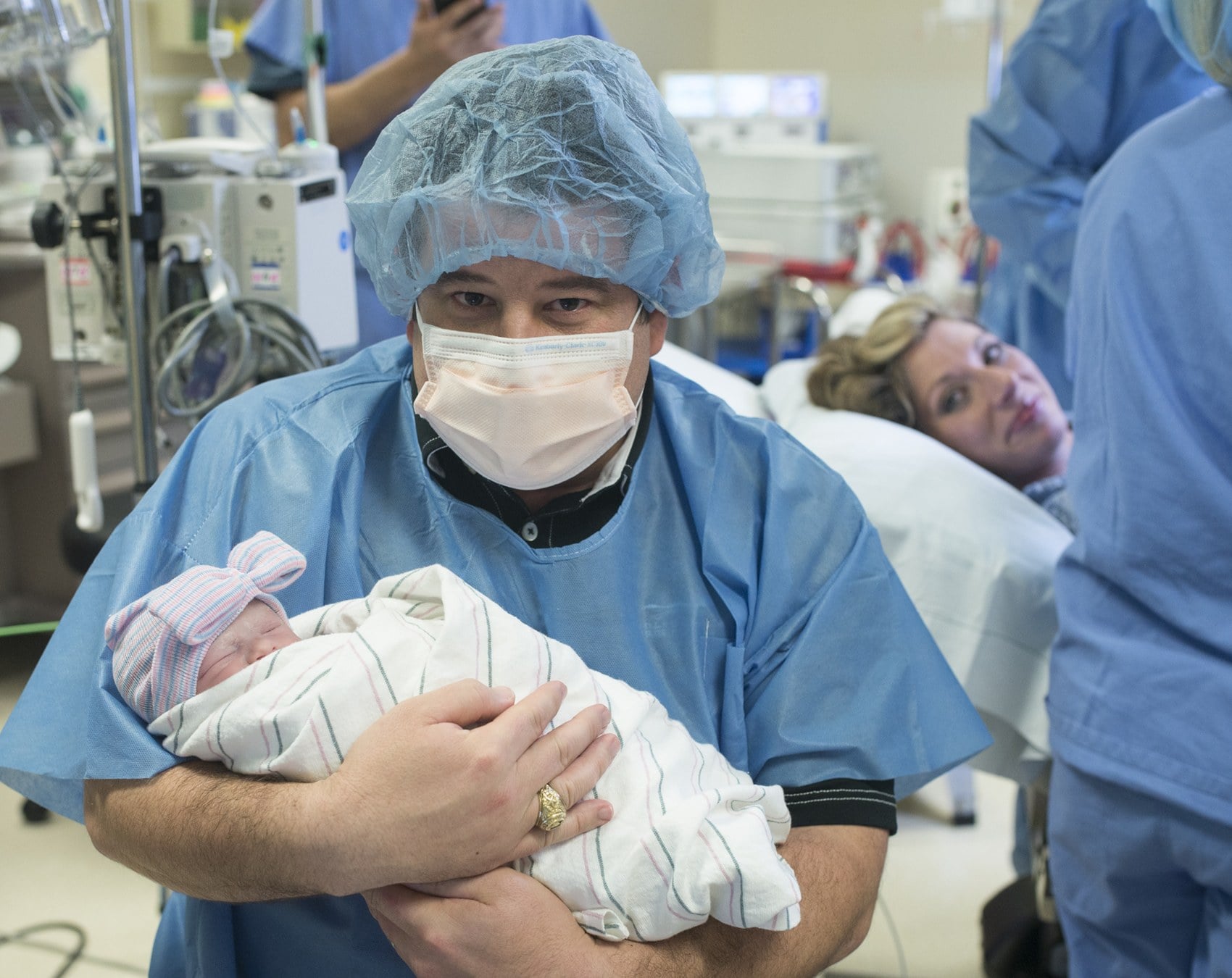 A father in surgical scrubs, cap and mask holds his newborn in the operating room while the mother watches from the table behind him.