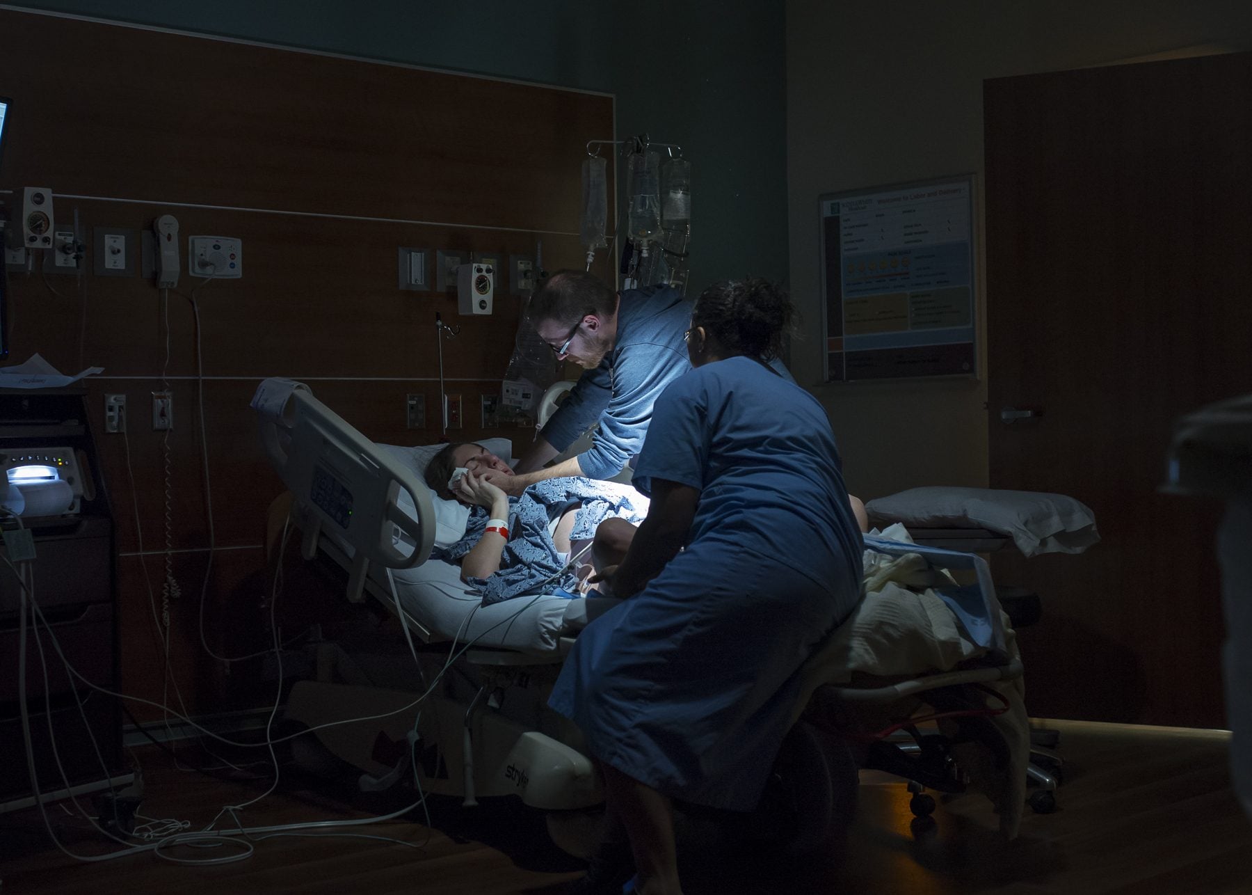 A darkened labor and delivery room at night. A nurse and the mother's partner support her at the bedside, lit only by a single lamp.