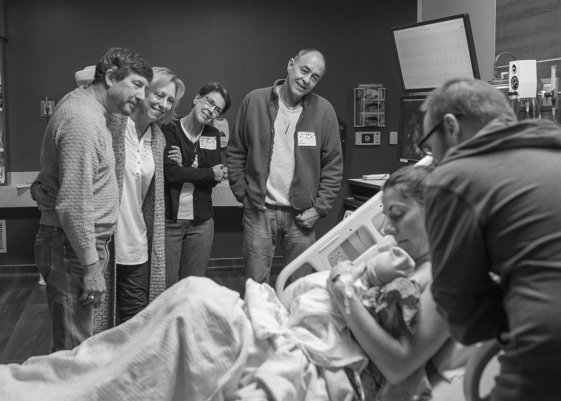 Black and white photograph of four grandparents standing along the wall of a delivery room, watching as the mother holds her newborn and the father leans in.