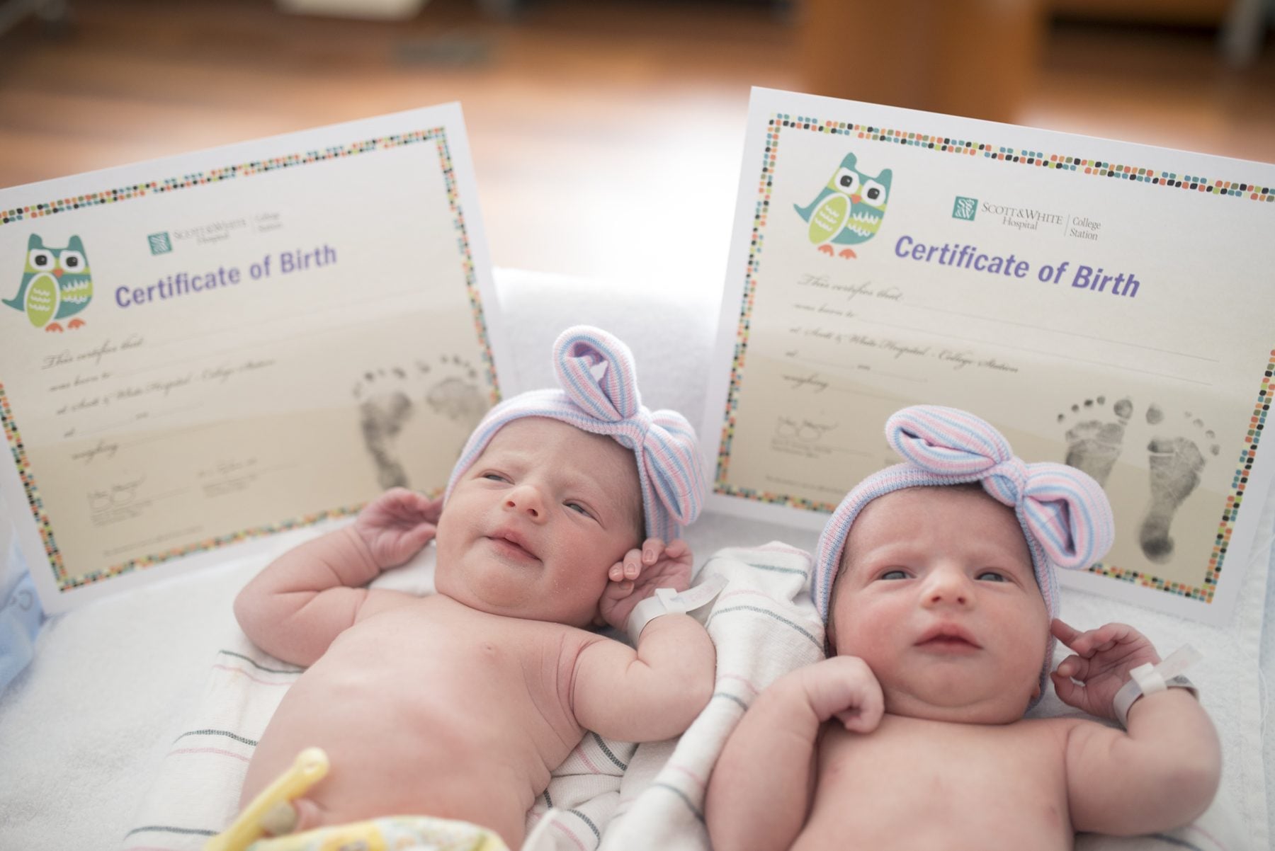 Newborn twins in striped hospital hats lying side by side in front of their two birth certificates.